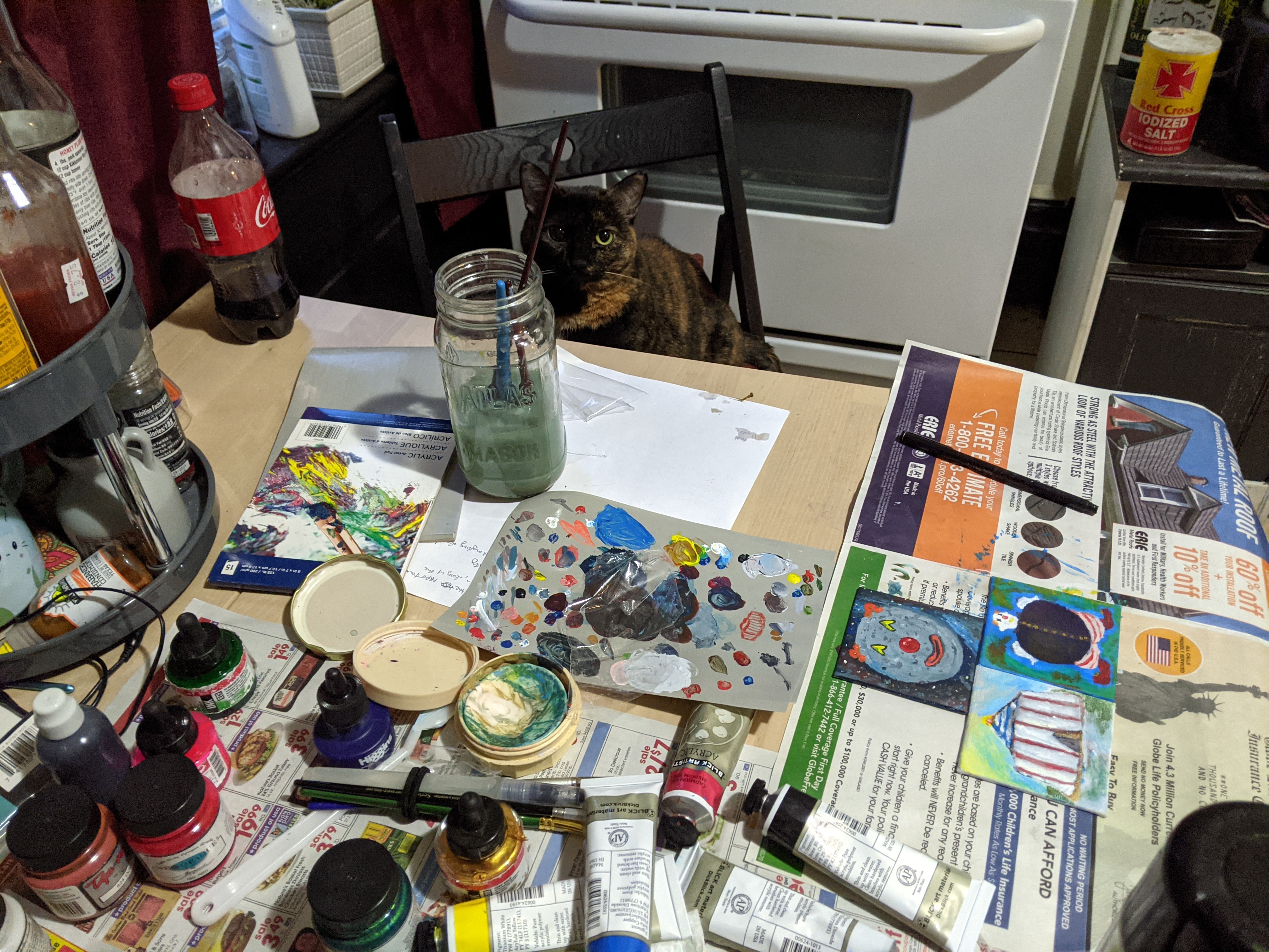 the same cat sitting at a desk that is absolutely covered in painting supplies